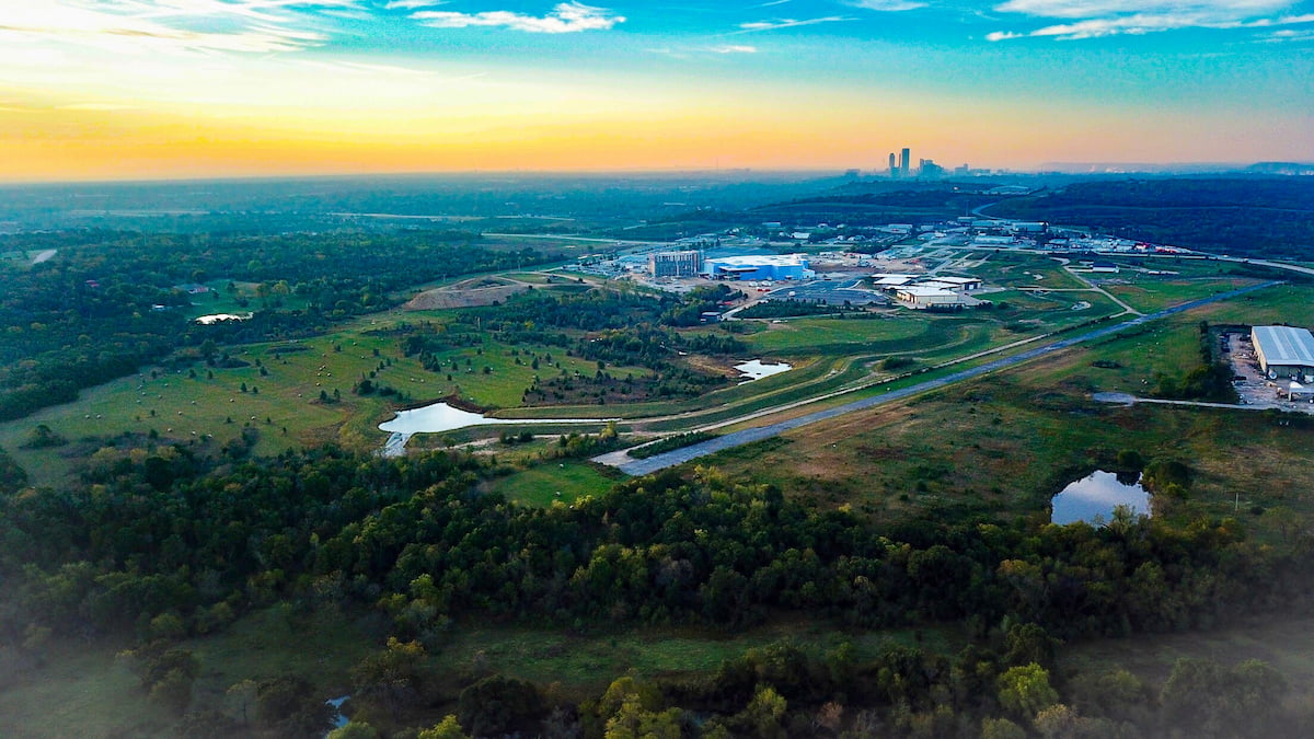 Drone photo from a very high altitude of a large flat landscape of grassy land, woodland, and industrial or office buildings. A runway cuts through the foreground and the skyline of Tulsa, Oklahoma rises in the distance. 